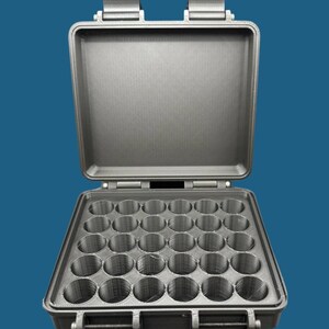 Puede incluir: Una caja de almacenamiento de plástico gris con una tapa con bisagras y una cuadrícula de 24 compartimentos redondos. La caja está abierta y vacía.