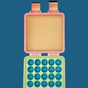 Puede incluir: Una caja de almacenamiento de plástico de color rosa pastel y verde con una tapa con bisagras y 25 compartimentos redondos. La caja está abierta, revelando los compartimentos vacíos.