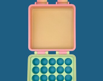 May include: A square, open storage case featuring a light pink lid and a light green base. The interior of the base contains a grid of twenty-five circular indentations in a bright blue color. Two hinges are visible at the top edge.