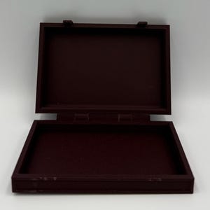 May include: An open, rectangular, burgundy-colored wooden box with a hinged lid. The box is empty and sits against a white background. The box is made of wood and has a simple, classic design.