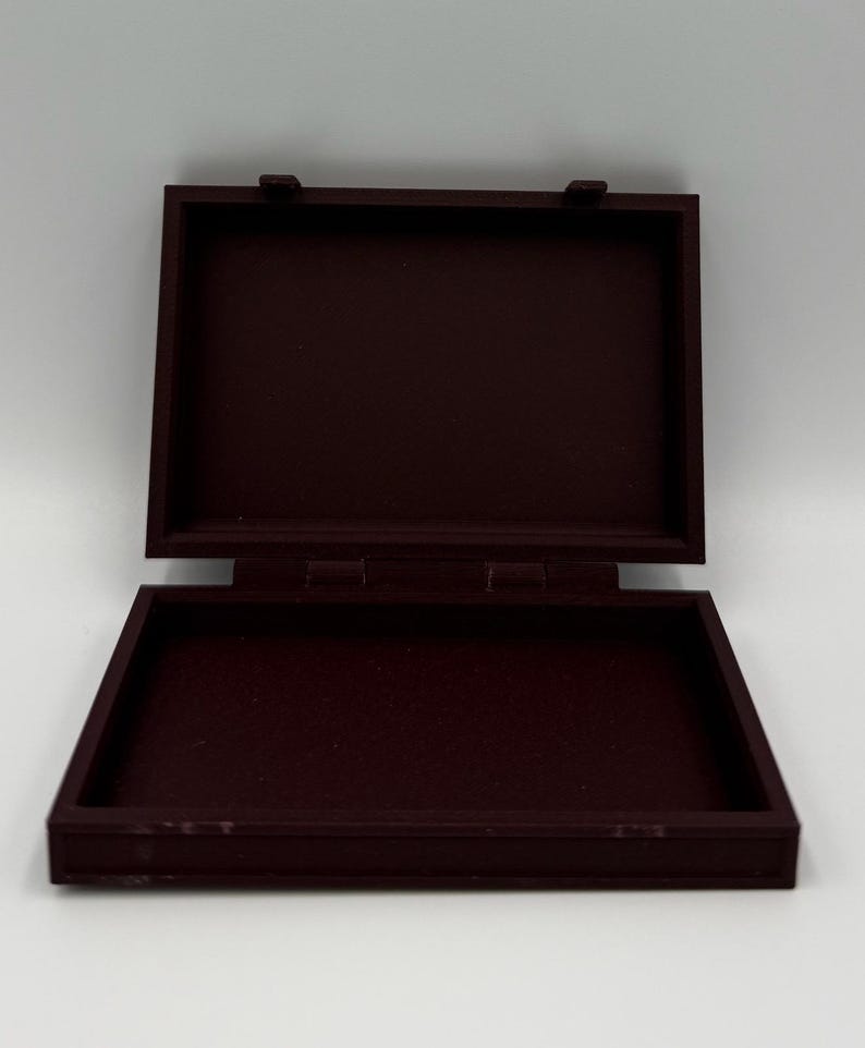 May include: An open, rectangular, burgundy-colored wooden box with a hinged lid. The box is empty and sits against a white background. The box is made of wood and has a simple, classic design.