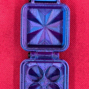 Puede incluir: Un recipiente cuadrado con bisagras, de color azul y morado iridiscente con acabado texturizado. El interior de la tapa presenta un patr&oacute;n de estrella radiante, mientras que la base tiene cuatro hendiduras circulares.