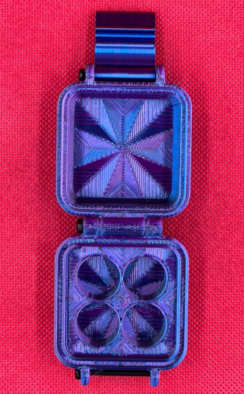 Puede incluir: Un recipiente cuadrado con bisagras, de color azul y morado iridiscente con acabado texturizado. El interior de la tapa presenta un patr&oacute;n de estrella radiante, mientras que la base tiene cuatro hendiduras circulares.
