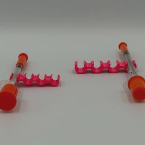 May include: Two medical syringes with orange caps and pink holders are displayed against a white background. The syringes are partially filled with a clear liquid. The holders have a wavy design.