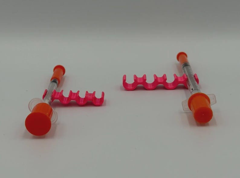 May include: Two medical syringes with orange caps and pink holders are displayed against a white background. The syringes are partially filled with a clear liquid. The holders have a wavy design.
