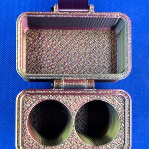 May include: A rectangular, hinged storage case with a metallic, multi-colored finish featuring a textured, cross-hatched pattern. The top section is an open compartment, while the bottom section contains two circular, hollow cylindrical openings.