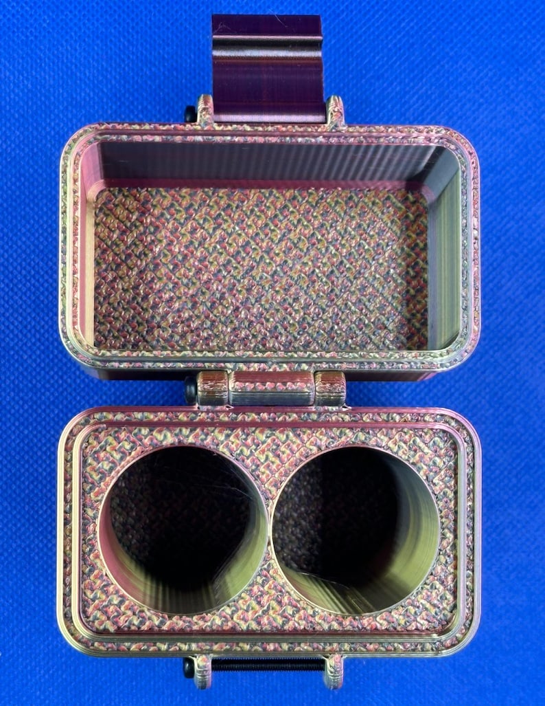 May include: A rectangular, hinged storage case with a metallic, multi-colored finish featuring a textured, cross-hatched pattern. The top section is an open compartment, while the bottom section contains two circular, hollow cylindrical openings.