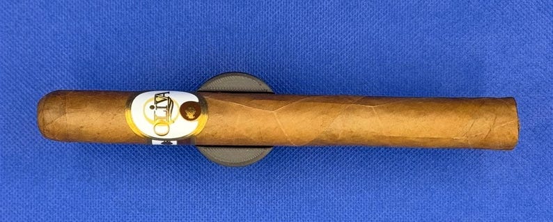 May include: A brown cigar with a white band that says "Oliva" on it. The cigar is resting on a black cigar rest.