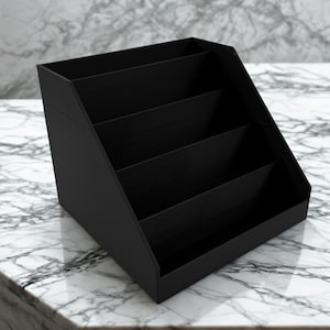 May include: A black, angled desk organizer with four compartments. The organizer is made of a solid material and sits on a white and gray marble surface. The compartments are open and ready to hold office supplies.