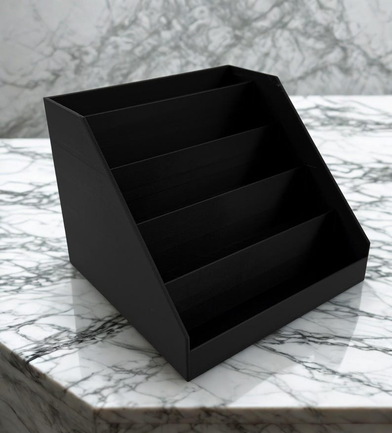 May include: A black, angled desk organizer with four compartments. The organizer is made of a solid material and sits on a white and gray marble surface. The compartments are open and ready to hold office supplies.
