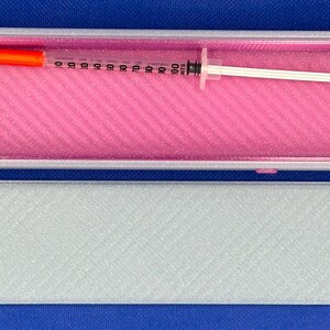 May include: A rectangular gray storage case with a bright pink interior lining. Inside the open case lies a clear plastic syringe featuring a bright orange cap on one end and a white plunger on the other, marked with black measurement units.