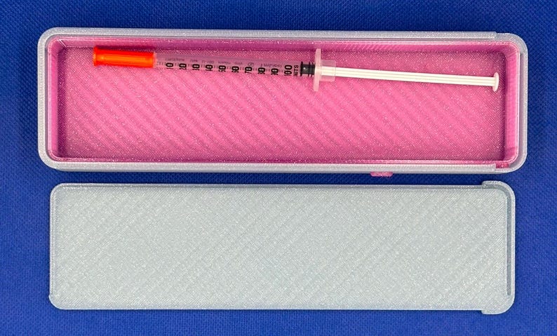 May include: A rectangular gray storage case with a bright pink interior lining. Inside the open case lies a clear plastic syringe featuring a bright orange cap on one end and a white plunger on the other, marked with black measurement units.