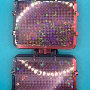 May include: A black, rectangular, hinged display case with two clear panels featuring a diagonal striped texture and a holographic star pattern. The case is connected by a central hinge and has a black tab at the top for hanging or mounting.