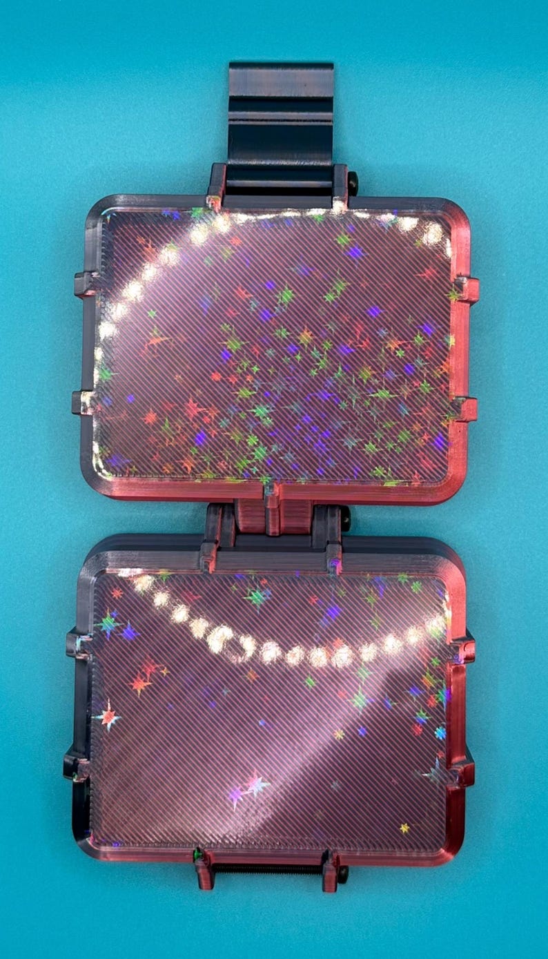 May include: A black, rectangular, hinged display case with two clear panels featuring a diagonal striped texture and a holographic star pattern. The case is connected by a central hinge and has a black tab at the top for hanging or mounting.