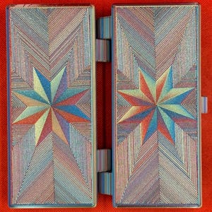 May include: A rectangular, hinged case featuring a vibrant, multi-colored star pattern on both panels. The surface displays a textured, iridescent finish with geometric lines radiating from the center of each star in shades of blue, red, and gold.
