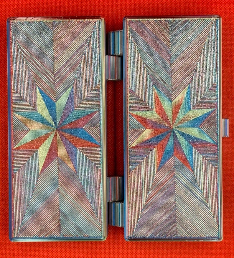 May include: A rectangular, hinged case featuring a vibrant, multi-colored star pattern on both panels. The surface displays a textured, iridescent finish with geometric lines radiating from the center of each star in shades of blue, red, and gold.