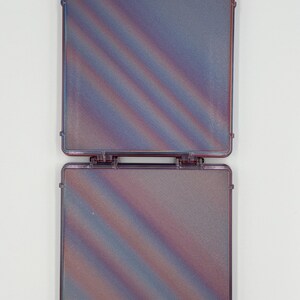 May include: A square, hinged, translucent purple plastic case with a textured surface featuring diagonal iridescent color shifts. The case has two small, plaid-patterned fabric tabs attached to the top edge for opening and closing.