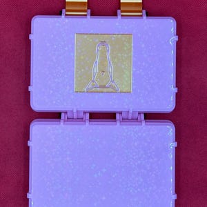 May include: Open, light purple, hinged case with a gold-colored emblem. The case is decorated with a subtle star pattern. Two brown and gold connectors are attached to the top of the case. The case is open, revealing the interior.