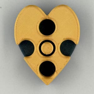 May include: A heart-shaped object with a gold-colored exterior and three black circular cutouts. The object has a textured surface and a geometric design, with a smaller circle in the center.