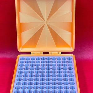 May include: A square orange storage case with a hinged lid featuring a sunburst pattern. The interior contains a light purple insert with a grid of one hundred circular holes for organizing small items. The case has two front latches.