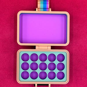 Puede incluir: Un estuche de almacenamiento rectangular de color melocot&oacute;n con tapa abatible. El interior presenta una secci&oacute;n superior morada s&oacute;lida y una secci&oacute;n inferior con quince hendiduras circulares moradas sobre una base verde claro. Una leng&uuml;eta a rayas arco&iacute;ris est&aacute; unida al borde superior.