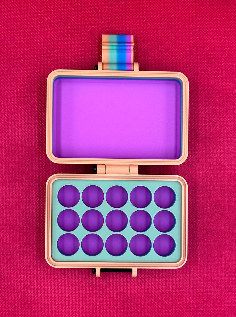 Puede incluir: Un estuche de almacenamiento rectangular de color melocot&oacute;n con tapa abatible. El interior presenta una secci&oacute;n superior morada s&oacute;lida y una secci&oacute;n inferior con quince hendiduras circulares moradas sobre una base verde claro. Una leng&uuml;eta a rayas arco&iacute;ris est&aacute; unida al borde superior.