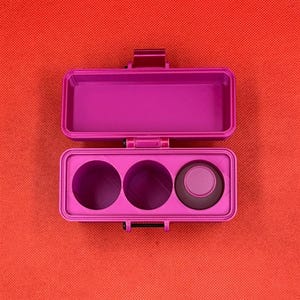 May include: A bright magenta rectangular storage case with a hinged lid. The interior features an open rectangular compartment on the top half and three circular recessed slots on the bottom half, with one small magenta cap inserted.