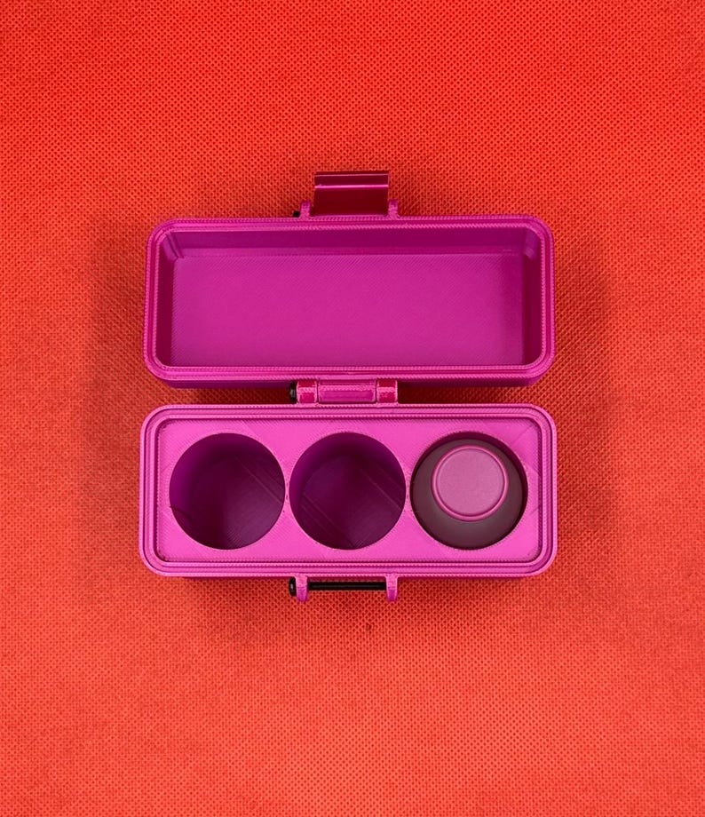 May include: A bright magenta rectangular storage case with a hinged lid. The interior features an open rectangular compartment on the top half and three circular recessed slots on the bottom half, with one small magenta cap inserted.