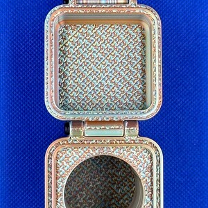 May include: A square, hinged metallic container with a copper-toned finish and a textured, repeating geometric pattern on its interior surfaces. The top section features a solid square base, while the bottom section has a circular opening.