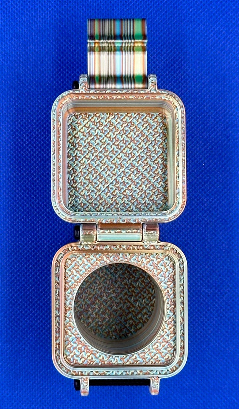 May include: A square, hinged metallic container with a copper-toned finish and a textured, repeating geometric pattern on its interior surfaces. The top section features a solid square base, while the bottom section has a circular opening.