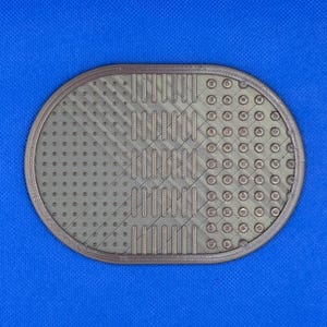 May include: An oval-shaped, metallic bronze-colored cleaning mat featuring a textured surface with three distinct sections: a grid of small raised dots, a series of vertical bars, and a pattern of larger circular indentations.