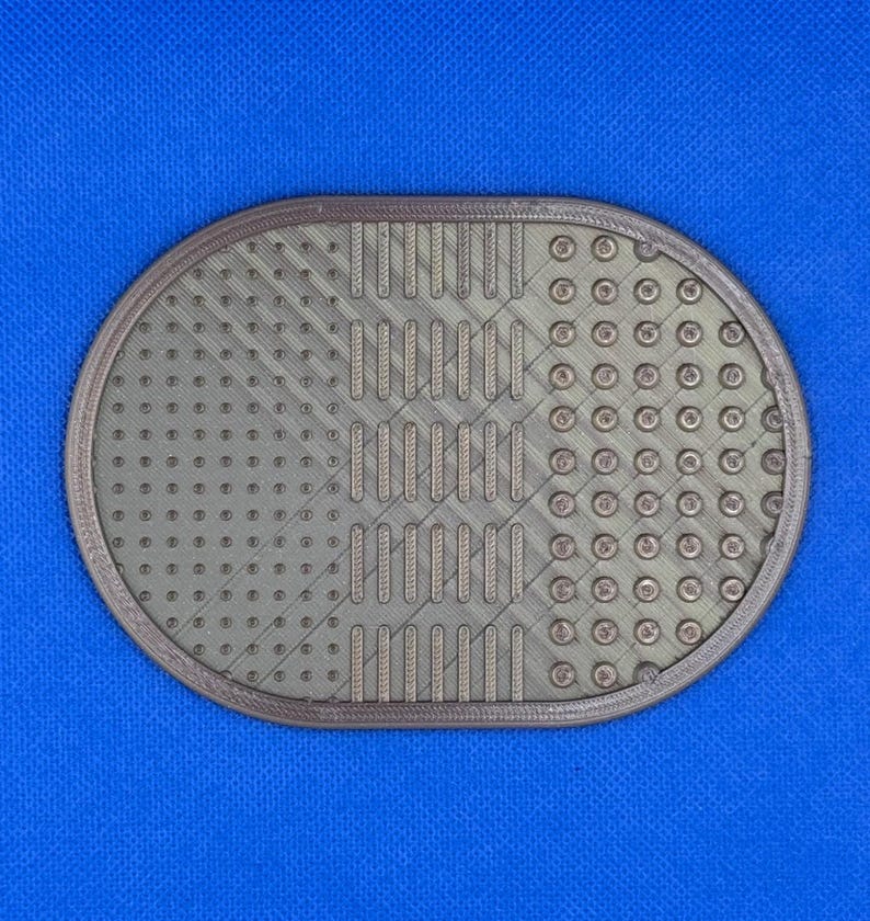 May include: An oval-shaped, metallic bronze-colored cleaning mat featuring a textured surface with three distinct sections: a grid of small raised dots, a series of vertical bars, and a pattern of larger circular indentations.