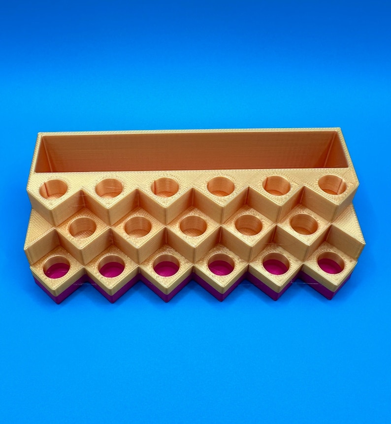 May include: A tiered, 3D-printed organizer featuring a rectangular storage compartment at the back and multiple circular slots arranged in a zigzag pattern. The structure is primarily gold-colored with a vibrant magenta base layer.