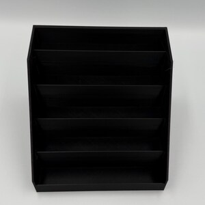 May include: A black, multi-compartment organizer with six horizontal shelves. The organizer is made of a solid, matte black material and is designed to hold and display various items. The design is simple and modern, suitable for home or office use.