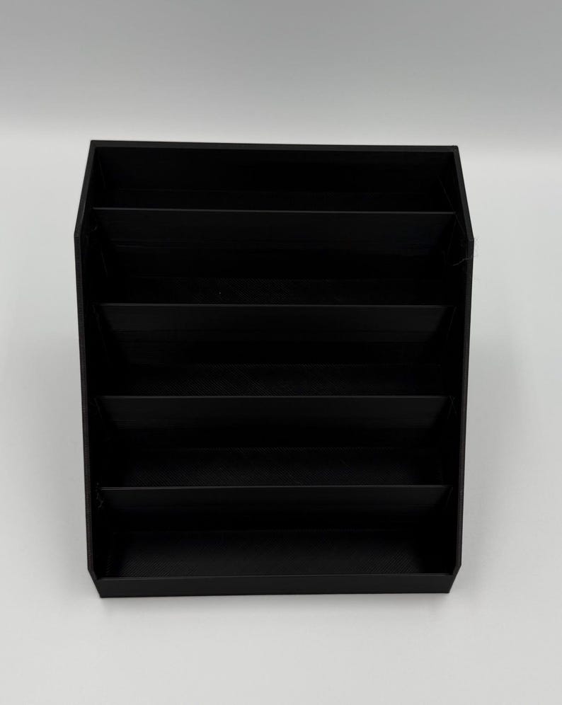May include: A black, multi-compartment organizer with six horizontal shelves. The organizer is made of a solid, matte black material and is designed to hold and display various items. The design is simple and modern, suitable for home or office use.