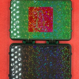 May include: Two open, small, rectangular cases with a glittery finish. The top case is green with a red square featuring a design. The bottom case is black with a similar glittery finish. Both cases have red accents and are set against a red background.