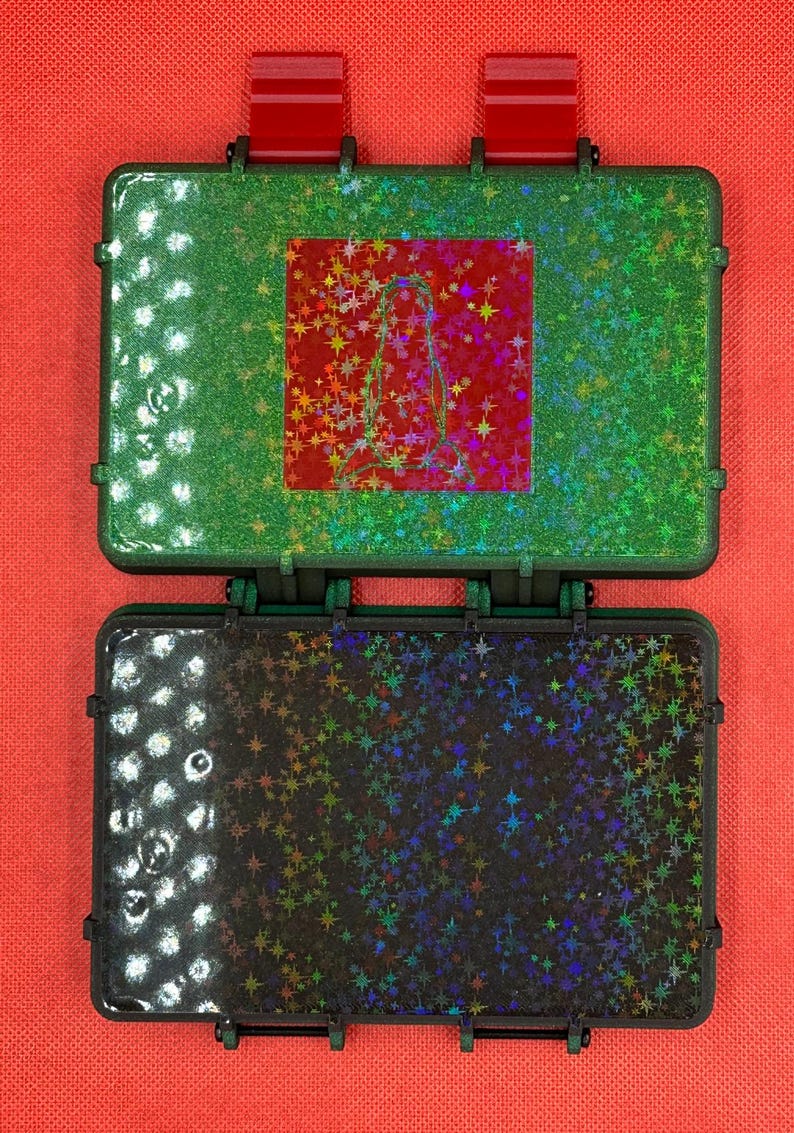 May include: Two open, small, rectangular cases with a glittery finish. The top case is green with a red square featuring a design. The bottom case is black with a similar glittery finish. Both cases have red accents and are set against a red background.