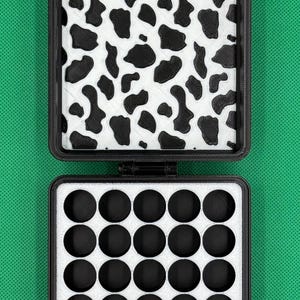 May include: A black square storage case with a hinged lid. The interior lid features a white and black cow print pattern. The base contains a white foam insert with twenty-five circular cutouts arranged in a five-by-five grid pattern.