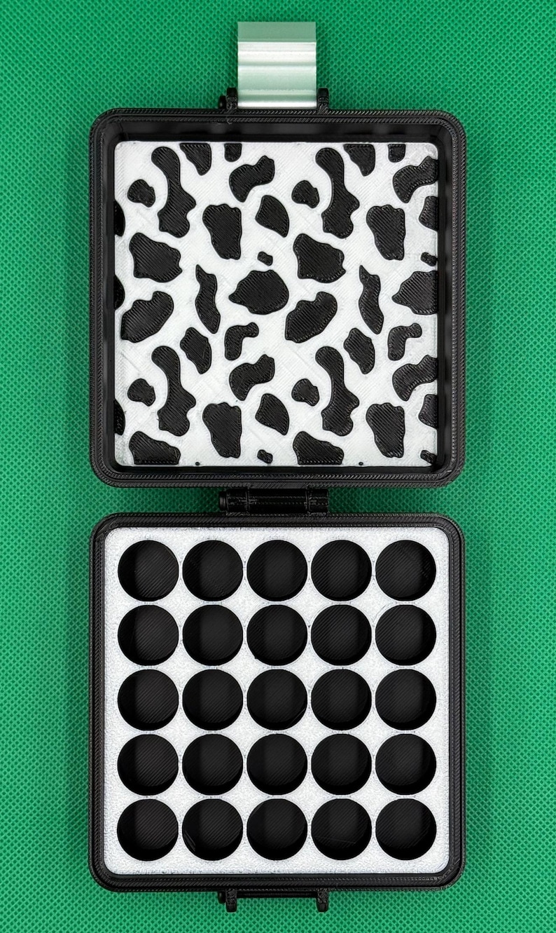 May include: A black square storage case with a hinged lid. The interior lid features a white and black cow print pattern. The base contains a white foam insert with twenty-five circular cutouts arranged in a five-by-five grid pattern.