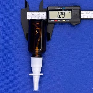 May include: A dark amber glass spray bottle with a white plastic pump nozzle. A black digital caliper is positioned across the bottle, displaying a measurement of 1.29 inches on its screen, indicating the diameter of the bottle neck.