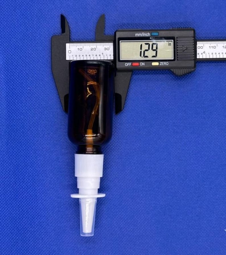 May include: A dark amber glass spray bottle with a white plastic pump nozzle. A black digital caliper is positioned across the bottle, displaying a measurement of 1.29 inches on its screen, indicating the diameter of the bottle neck.