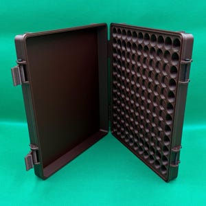 May include: A brown plastic storage case with a hinged lid. The case has a grid of round compartments on one side and a smooth surface on the other side.