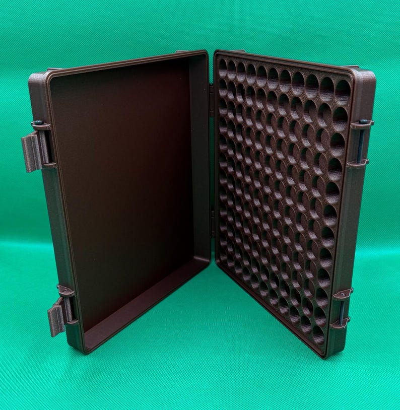 May include: A brown plastic storage case with a hinged lid. The case has a grid of round compartments on one side and a smooth surface on the other side.