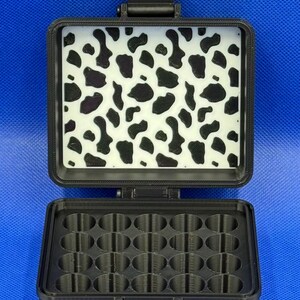 May include: A black plastic storage case with a hinged lid. The lid has a white and black cow print design. The case has 20 round compartments.