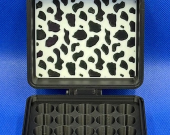 May include: A black rectangular storage case with a hinged lid. The interior of the lid features a white panel with a black cow print pattern. The base of the case contains a grid of sixteen circular compartments for organizing small items.