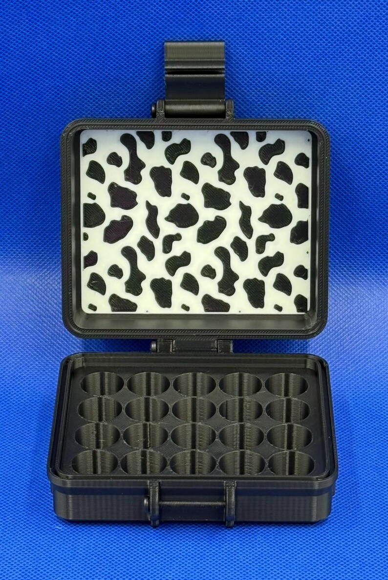 May include: A black rectangular storage case with a hinged lid. The interior of the lid features a white panel with a black cow print pattern. The base of the case contains a grid of sixteen circular compartments for organising small items.