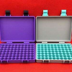 May include: Two rectangular storage cases with hinged lids. The left case is purple with black latches and a purple interior grid. The right case is gray with light blue latches and a light blue interior grid, each featuring multiple circular slots.