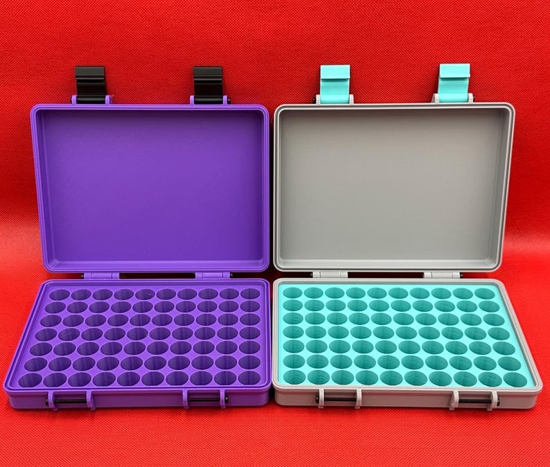 May include: Two rectangular storage cases with hinged lids. The left case is purple with black latches and a purple interior grid. The right case is gray with light blue latches and a light blue interior grid, each featuring multiple circular slots.