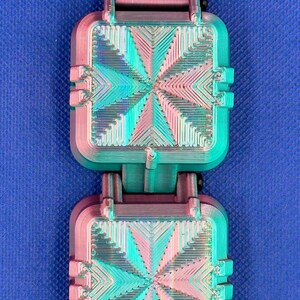 May include: A pair of square, 3D-printed components featuring a gradient color shift from magenta to teal. Each piece displays a geometric, radiating line pattern on its surface and is connected by a dark purple, ribbed strap segment.