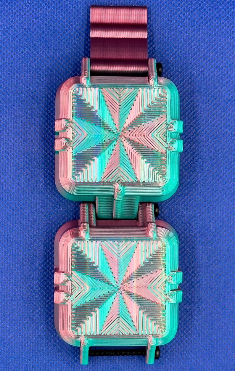 May include: A pair of square, 3D-printed components featuring a gradient color shift from magenta to teal. Each piece displays a geometric, radiating line pattern on its surface and is connected by a dark purple, ribbed strap segment.